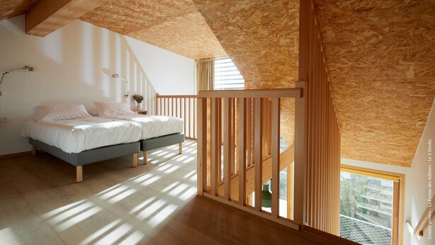 Ein Schlafzimmer mit schrägen Wänden, Holzbalken, einem Bett mit weißer Bettwäsche, einem Nachttisch und einem Fenster mit Blick auf Grünflächen.