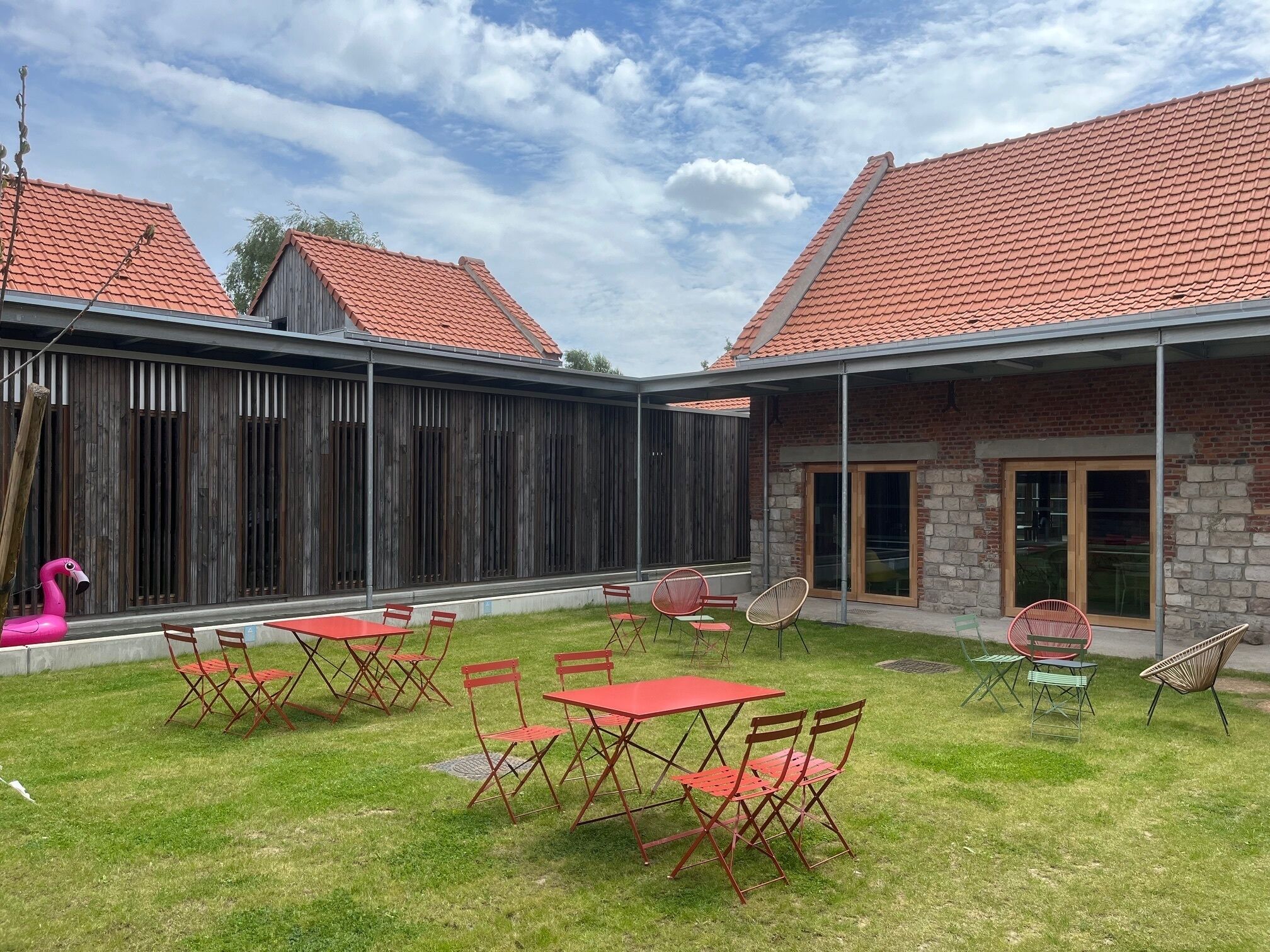 Un patio con tejado de tejas rojas, cerca de madera y zona ajardinada con mesas y sillas rojas.