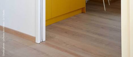 Una habitación con piso de madera, un armario amarillo con manijas negras, una silla blanca y una pequeña mesa con un cuenco.