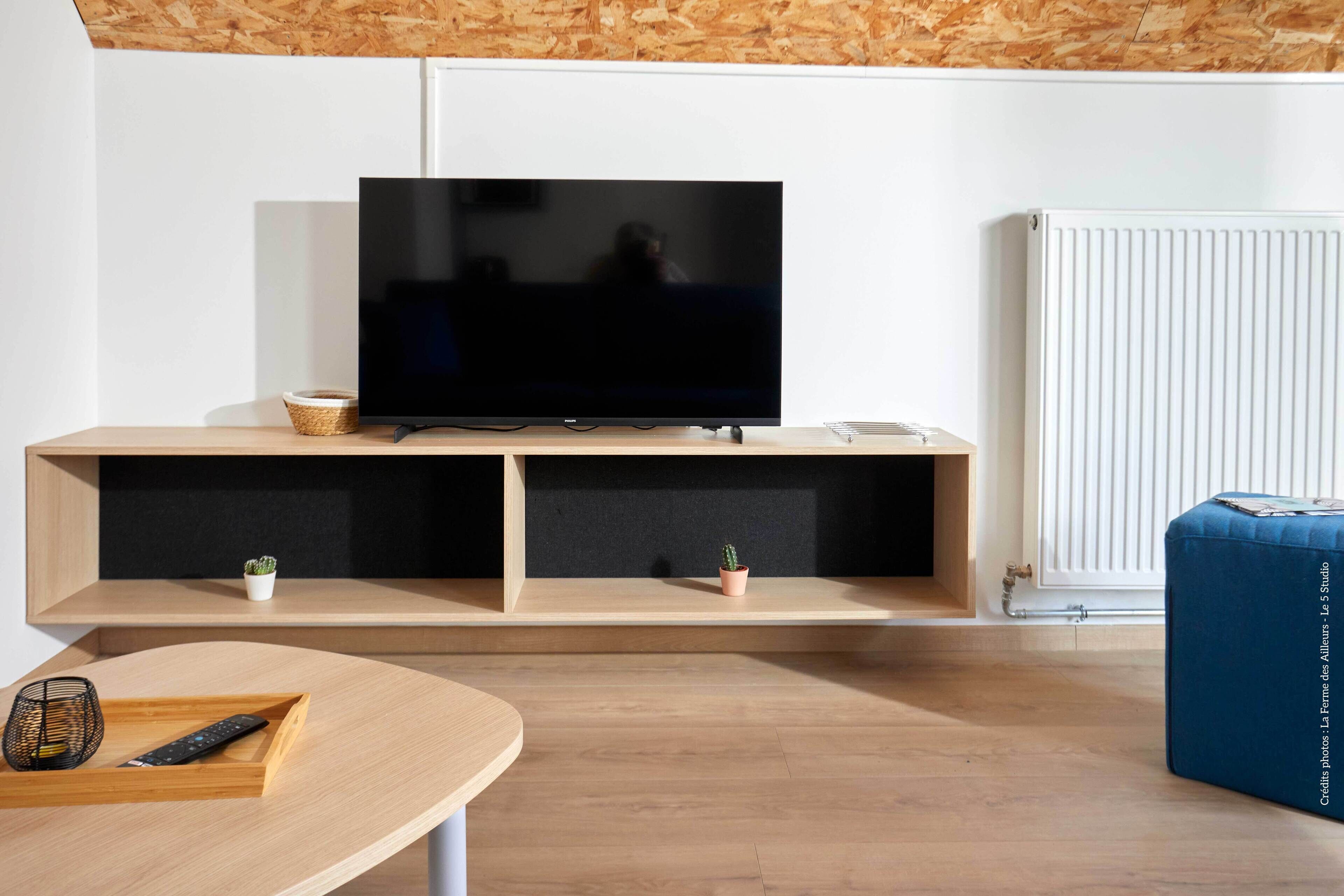 A modern living room with a flat-screen TV on a wooden stand, a small table with a remote control, and a blue ottoman.