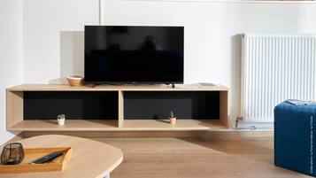 A modern living room with a flat-screen TV on a wooden stand, a small table with a remote control, and a blue ottoman.