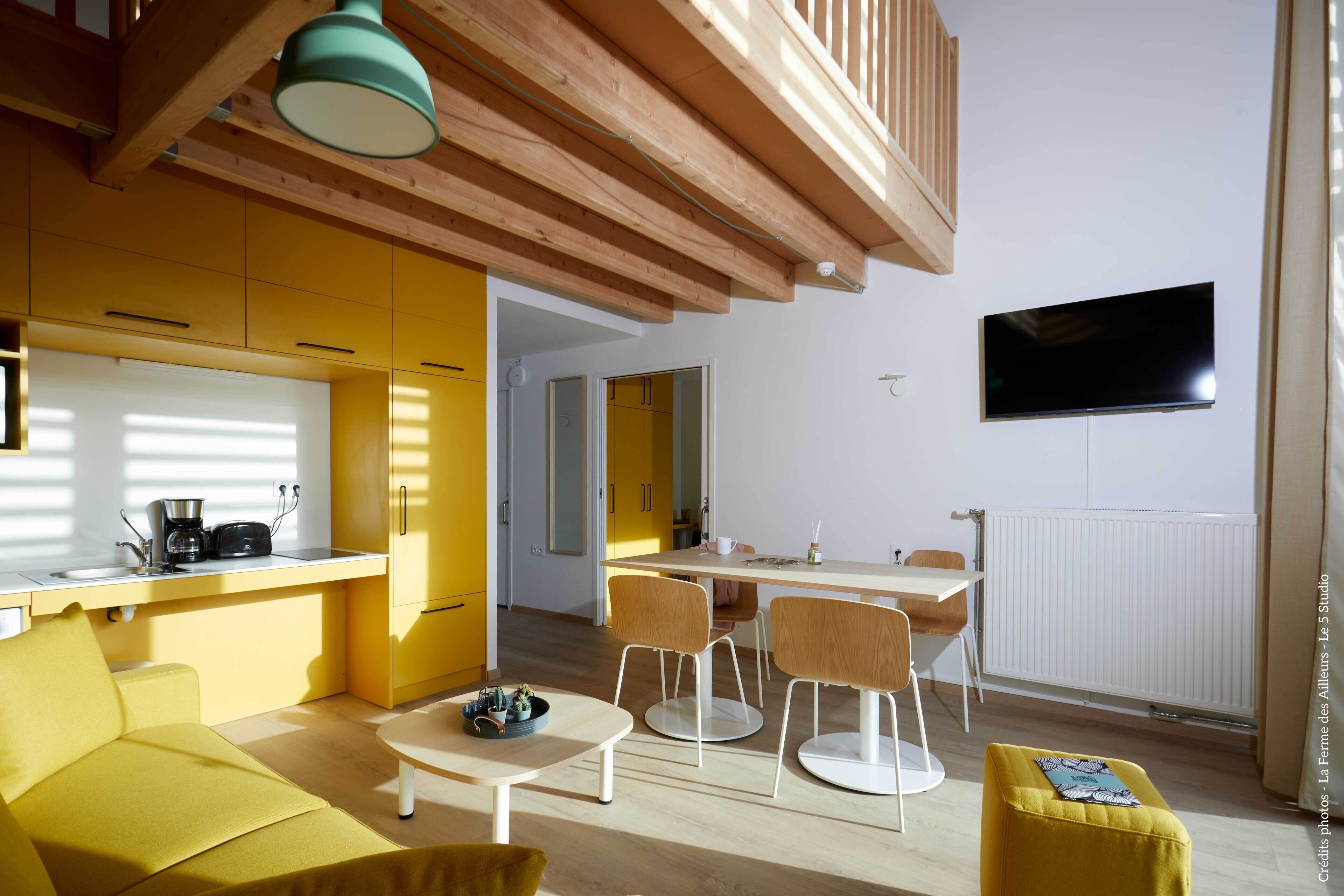 Een moderne woonkamer met gele wanden, een houten plafond, een flatscreen televisie aan de wand en een eethoek met een tafel en stoelen.