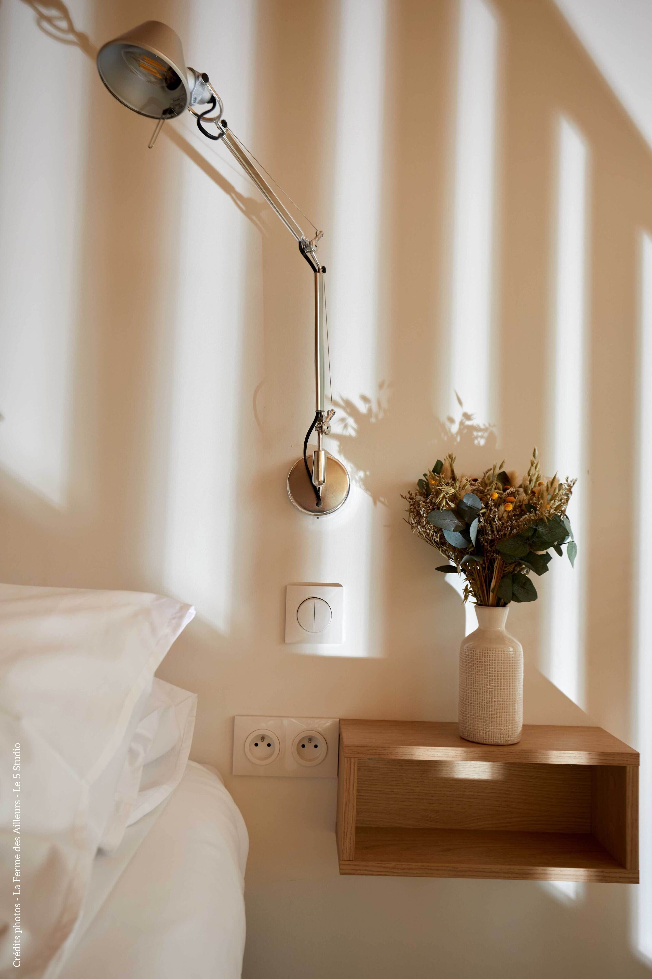 Ein ordentlich eingerichtetes Schlafzimmer mit Nachttischlampe, einer Vase mit Trockenblumen und einer Steckdose an der Wand.