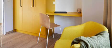 A room with a yellow wardrobe, a wooden chair, a yellow sofa, and a small table with a lamp.