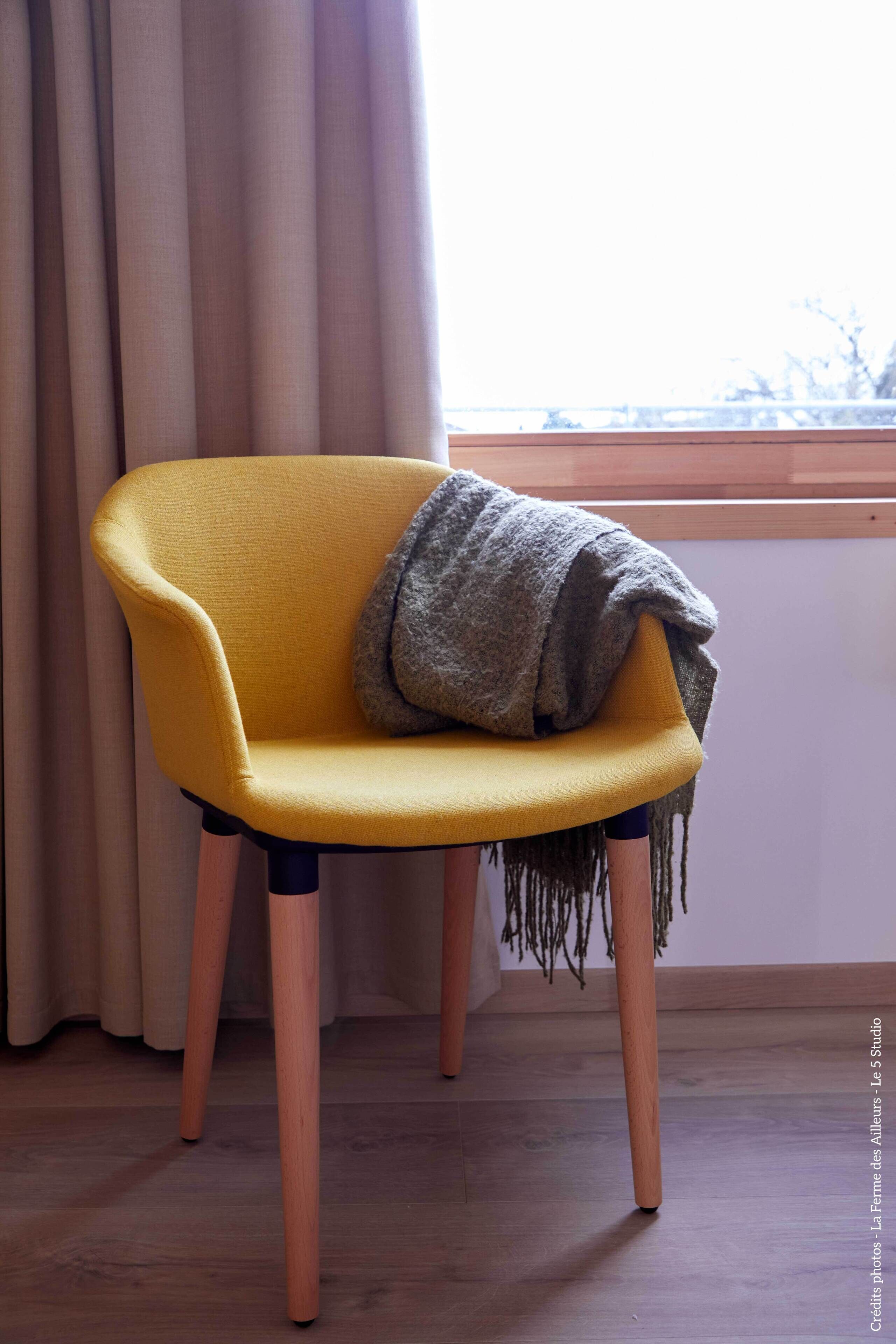 Ein gelber Stuhl mit einer grauen Decke, Holzbeinen und einem Fenster mit Vorhängen im Hintergrund.