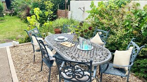 Een ronde metalen tafel met vier stoelen, omgeven door een tuin met verschillende planten en een witte muur op de achtergrond.
