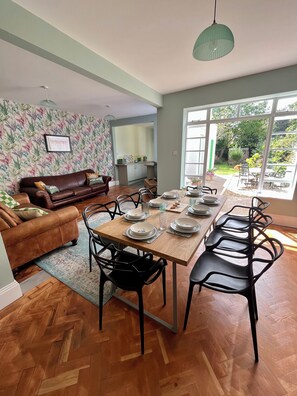 Un coin repas avec une table en bois dressée pour quatre personnes, entourée de chaises noires, d’un canapé en cuir marron et d’un papier peint à motifs.