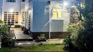Een huis met twee verdiepingen, met een witte gevel, een zichtbare schoorsteen en een kleine tuin met planten en een vogelhuisje.