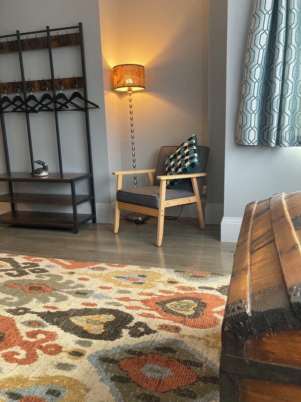 Une pièce comprenant une chaise en bois, un tapis à motifs, une lampe fixée au mur et un porte-manteau avec des cintres.
