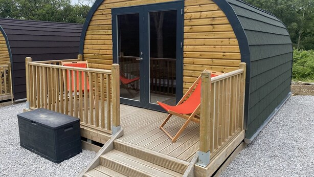 A wooden cabin with a curved roof, a glass door, and a small deck with a chair. - Sky Pod 1 (Pet Friendly) (Neath)