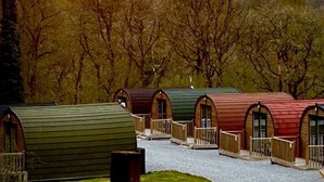 A row of log cabin-style cabins with arched doorways and wooden railings, set against a backdrop of trees. - Sky Pod 1 (Pet Friendly) (Neath)
