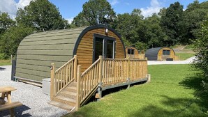 A series of wooden cabins with a deck and a covered porch, surrounded by a grassy area and trees. - Forest Pod 4 (Pet Friendly) -  a glamping pod that sleeps 2 guests  in 1 bedroom (Neath)