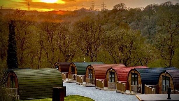 A row of unique, arched-roofed cabins with a gravel pathway leading to them, set against a backdrop of trees and a sunset. - A glamping pod that sleeps 2 guests  in 1 bedroom (Neath)