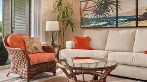 A living room with a wicker chair, a glass coffee table, a white sofa, and a large painting of a beach scene.