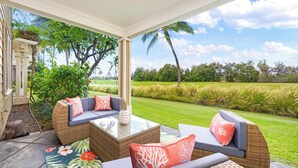 A covered outdoor seating area with wicker furniture and cushions, overlooking a lush green landscape and palm trees.