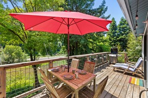 A wooden deck with a table and chairs, a red umbrella, and a grill. - Moondance - Contemporary Riverfront Home (Monte Rio)