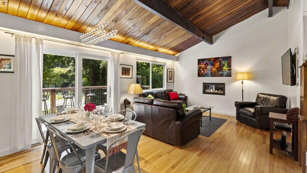 A spacious living area with a wooden ceiling, a fireplace, a dining table set for four, and a leather sofa. - Moondance - Contemporary Riverfront Home (Monte Rio)