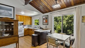 A modern kitchen with wooden cabinets, a central island, and a dining area with a view of greenery. - Moondance - Contemporary Riverfront Home (Monte Rio)