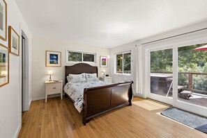 A bedroom with a wooden bed, two bedside tables with lamps, and large windows offering a view of a deck and trees. - Moondance - Contemporary Riverfront Home (Monte Rio)