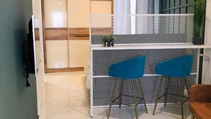 A room with a sliding door, a white partition with a mesh design, two blue chairs, a wooden bench, and a potted plant. - Lovely studio apart in Msida (L-MSIDA)