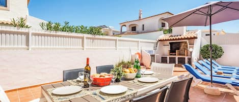 An outdoor dining area with a table set for four, surrounded by chairs, and a barbecue grill in the background.