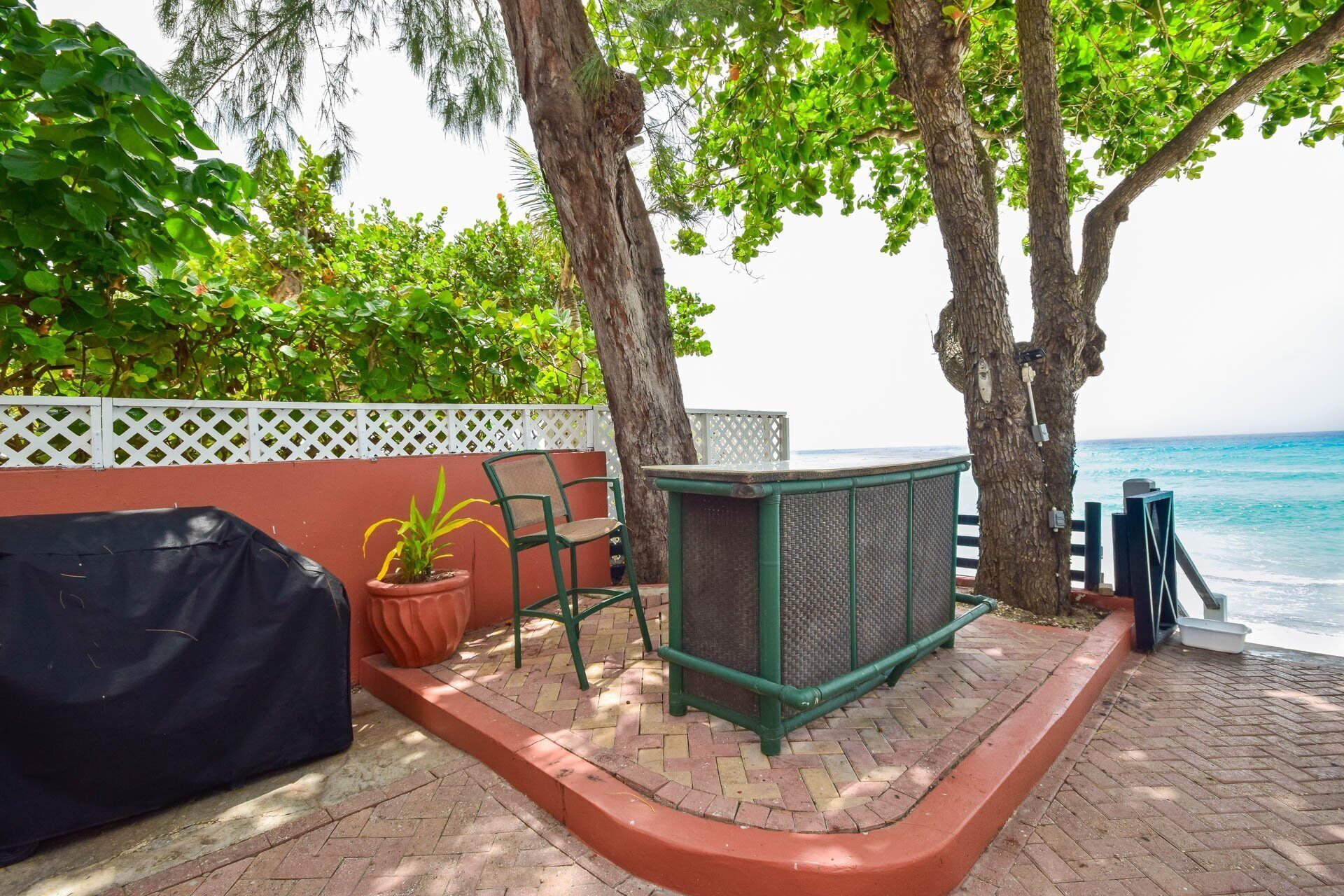 A patio with a grill, a chair, and a table near a white fence and trees.