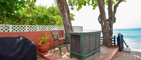 A patio with a grill, a chair, and a table near a white fence and trees.