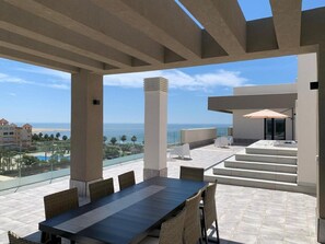 Un amplio espacio al aire libre para comer con vista al océano y a la playa, que incluye una mesa larga y sillas de mimbre.