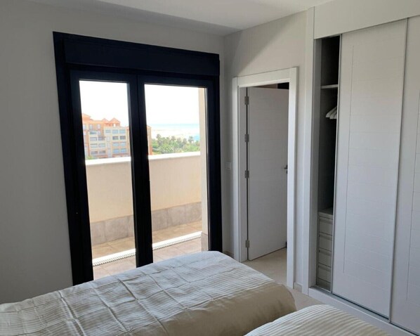 Un dormitorio con cama, armario y puerta corrediza que da a un balcón con vistas a edificios y a la playa.