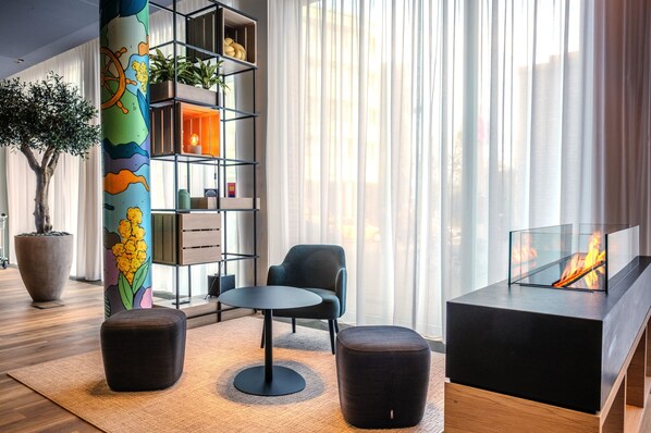 A modern living room with a round table, two ottomans, a bookshelf, and a fireplace. - IntercityHotel Lübeck (Lübeck)