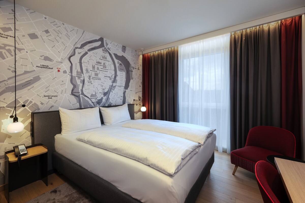 A hotel room with a large bed, a map on the wall, a desk with a chair, and a window with curtains.