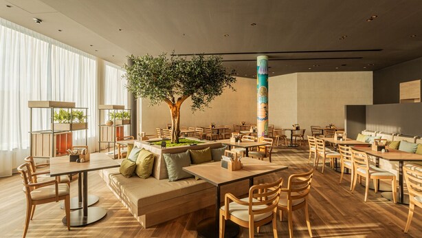 Un restaurant moderne avec des meubles en bois, un arbre central et une grande colonne ornée d’œuvres d’art.