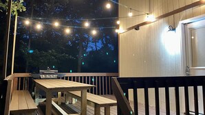 A wooden deck with a table and benches, string lights, and a grill.