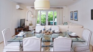 A dining area with a glass table, white chairs, and a sofa in the background. - Palisades 6a 3 Bedroom Townhouse (Oistins)