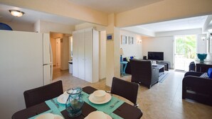 A dining area with a table set for four, a kitchen with a refrigerator, and a living area with a sofa and TV. - Bella Figura by BSL Rentals (Rockley)