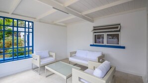 A room with a white sofa, a white armchair, a glass coffee table, a blue-framed window, and an air conditioning unit on the wall. - Bella Figura by BSL Rentals (Rockley)