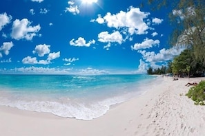 A beach with clear blue water, white sand, and scattered clouds.
