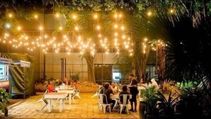 An outdoor dining area with tables and chairs, string lights overhead, and a building in the background. - Bella Figura by BSL Rentals (Rockley)