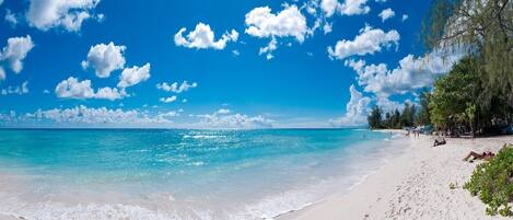 A beach with clear turquoise water, white sand, and scattered clouds.