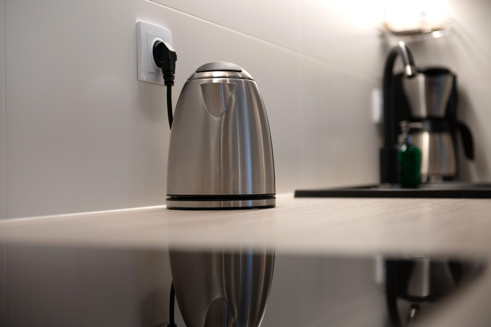 A stainless steel kettle on a reflective countertop with a power outlet and a coffee maker in the background.