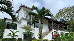 Una casa de dos pisos con balcón, rodeada de exuberante vegetación y palmeras.