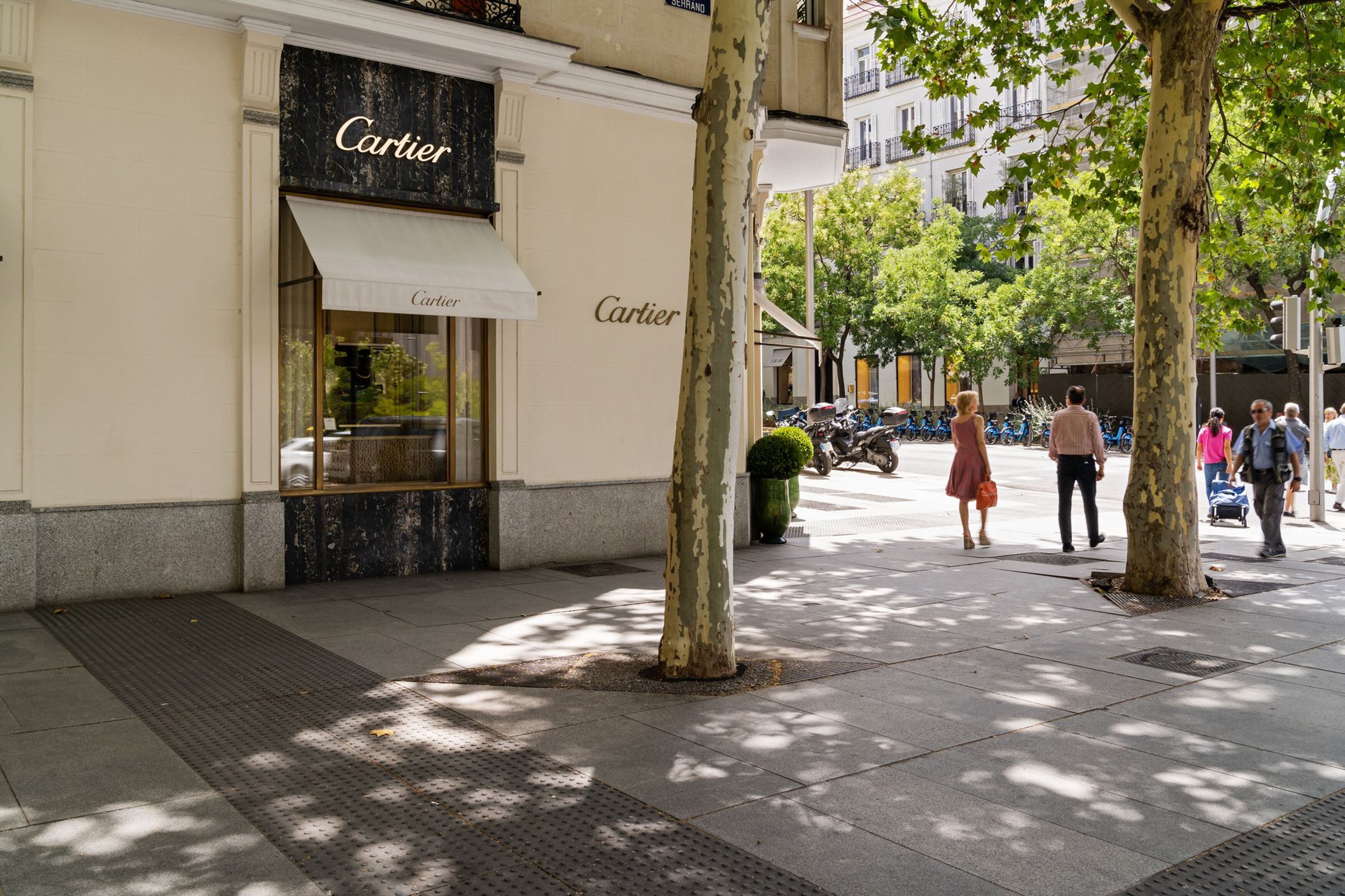 Eine Schaufensterfront mit dem deutlich erkennbaren Schriftzug 'Cartier', ein Bürgersteig und Bäume, die die Straße säumen.