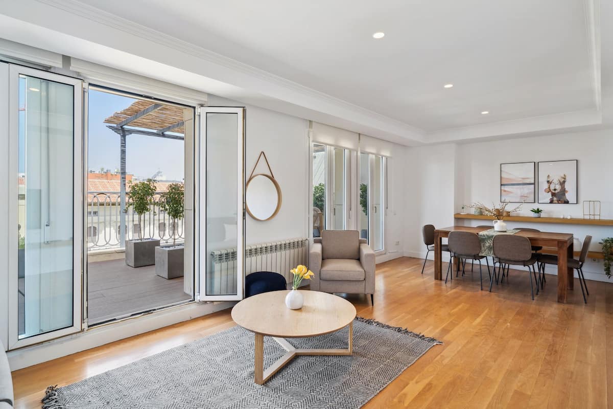 A spacious living area with a sliding glass door leading to a balcony, a round wooden table, a beige sofa, and a dining area with a wooden table and chairs.