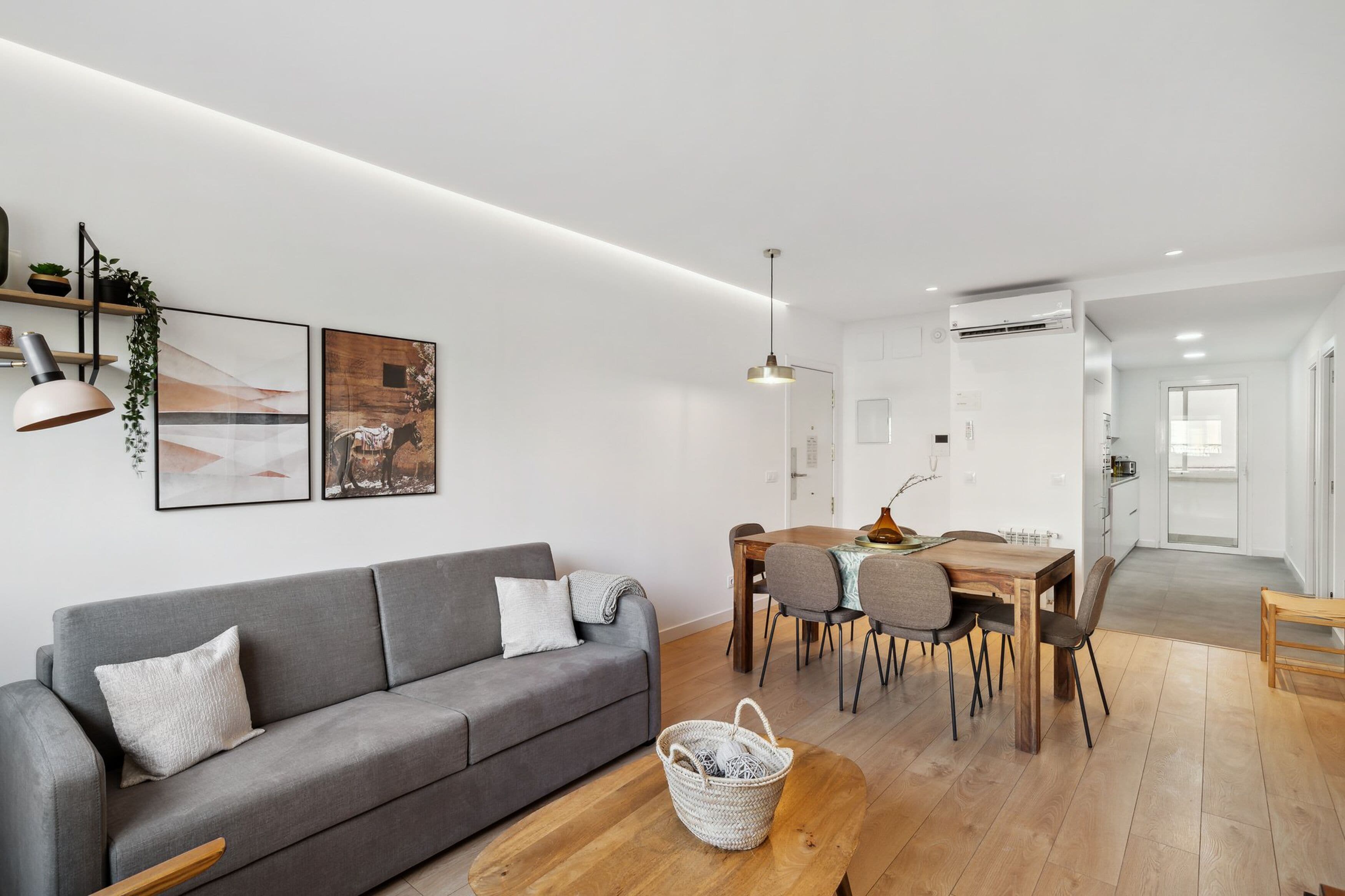 A modern living room with a grey sofa, wooden dining table, and chairs. There is a basket on the floor and a hanging pendant light.