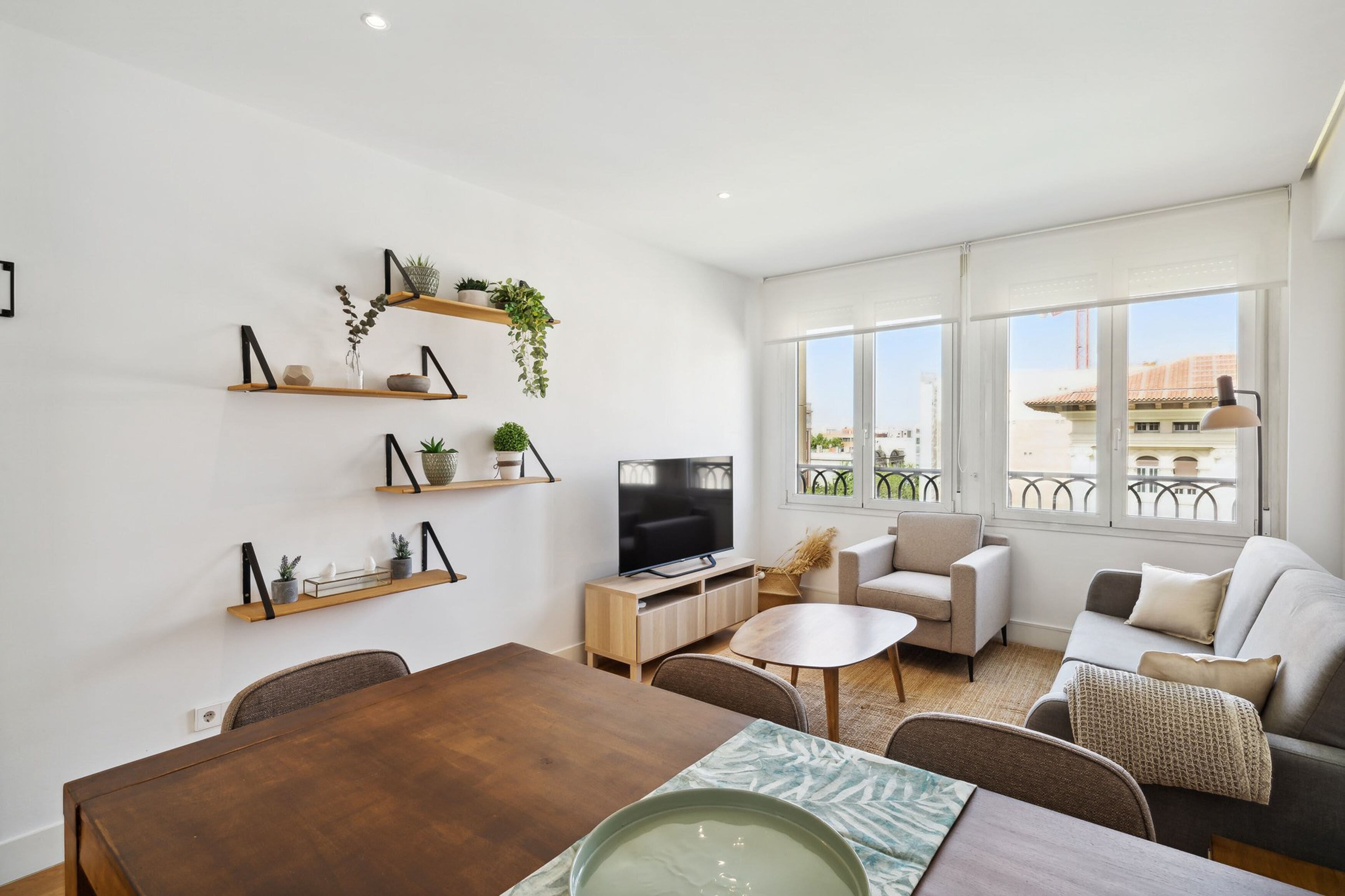 A modern living room with a wooden dining table, sofas, a TV, and shelves with plants.