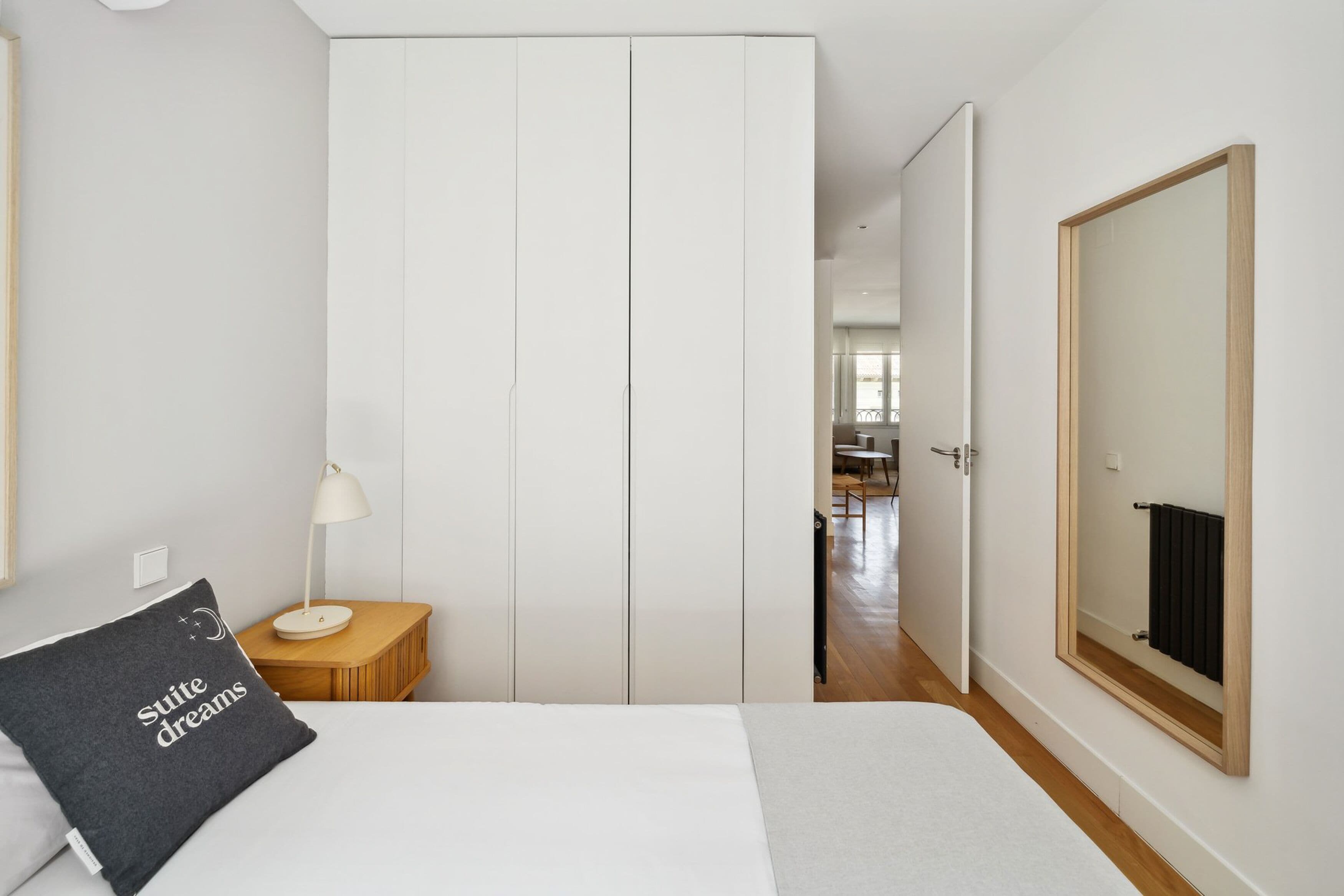 A bedroom with a bed, a bedside table with a lamp, a wardrobe, and a mirror on the wall.