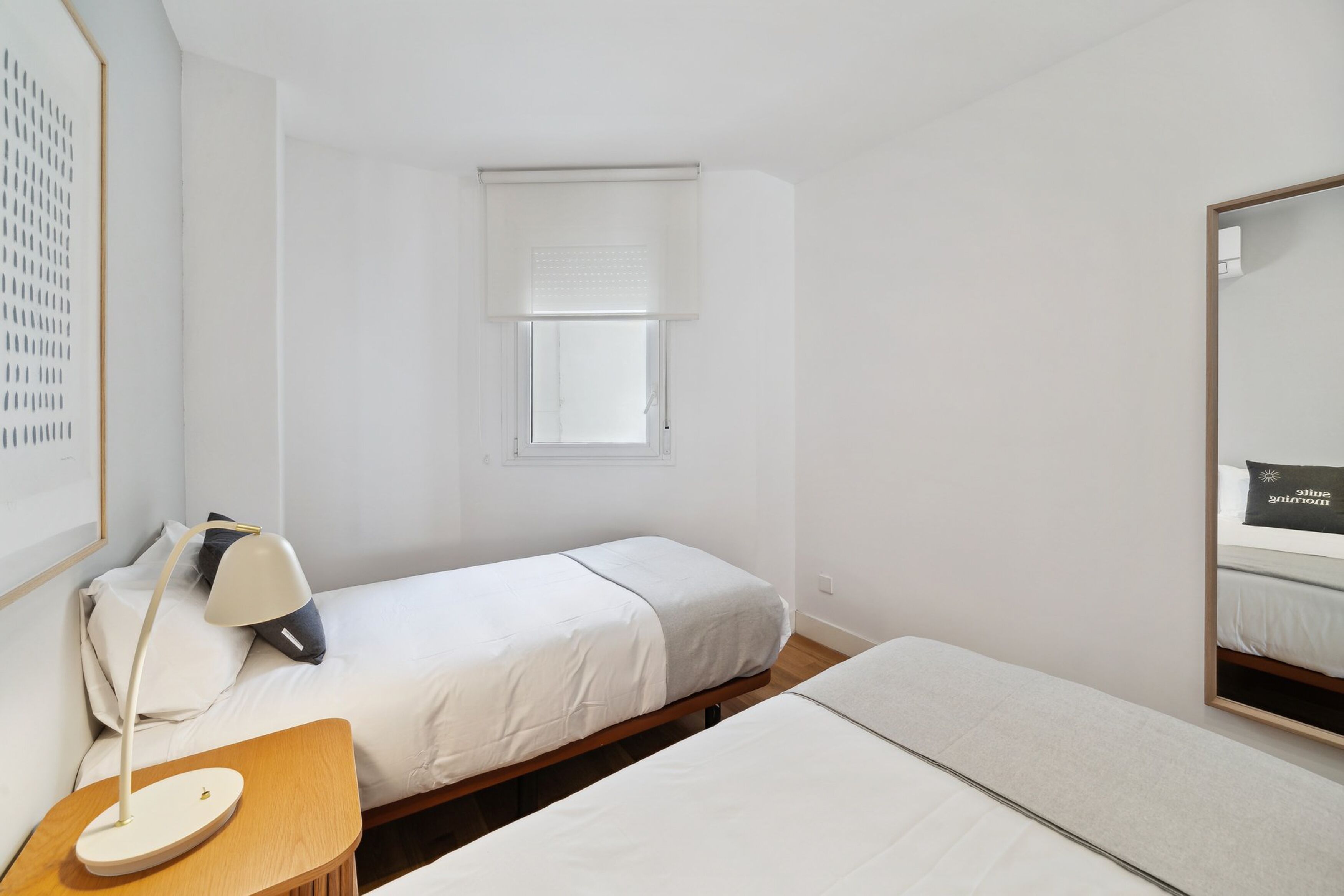 Habitación de hotel minimalista con dos camas, una mesita de noche pequeña con lámpara, una ventana con persianas y un espejo en la pared.