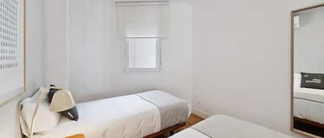 Habitación de hotel minimalista con dos camas, una mesita de noche pequeña con lámpara, una ventana con persianas y un espejo en la pared.