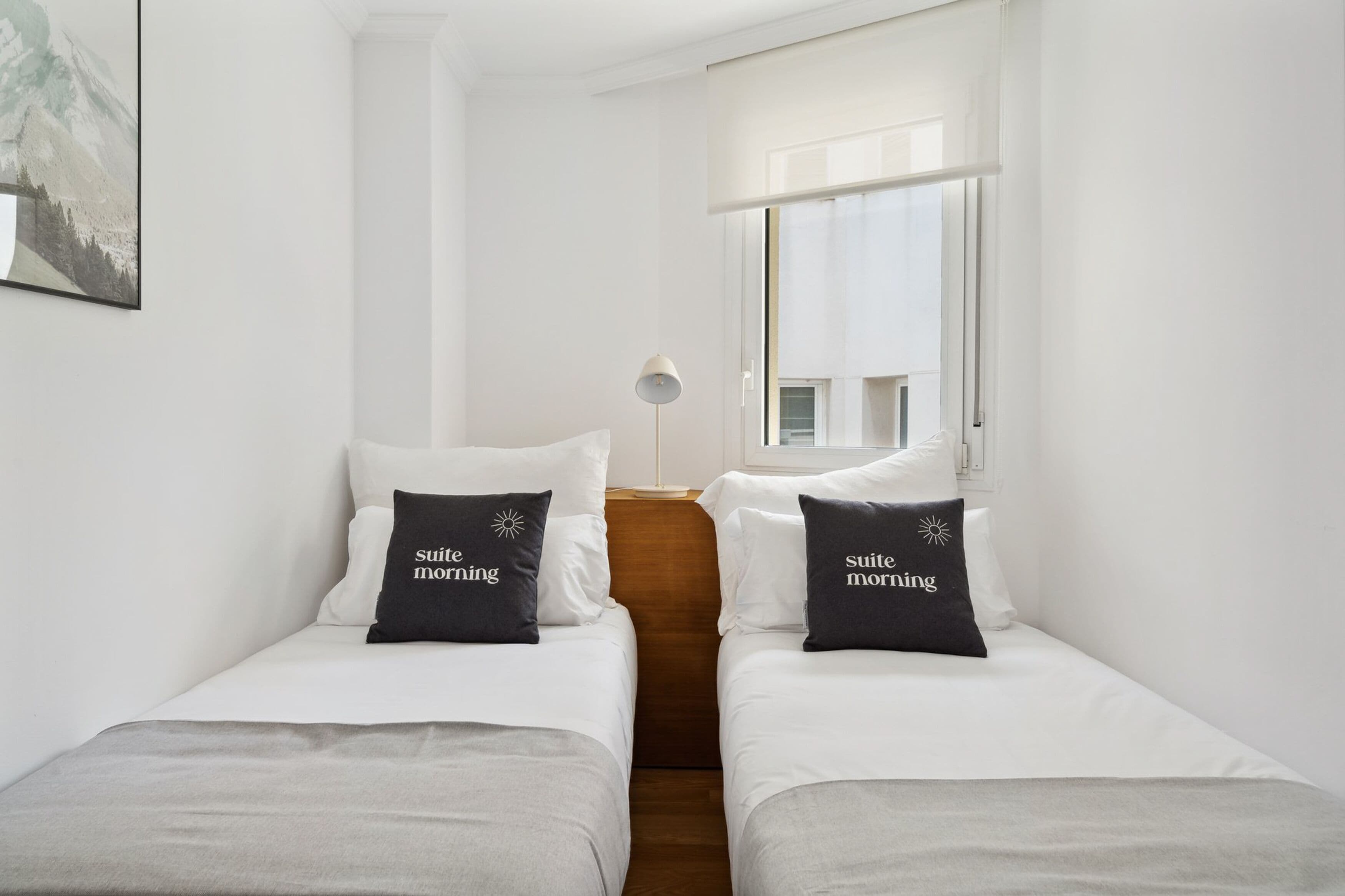 A hotel room with two single beds, each with a pillow that reads "suite morning." There is a window with blinds, a floor lamp, and a painting on the wall.