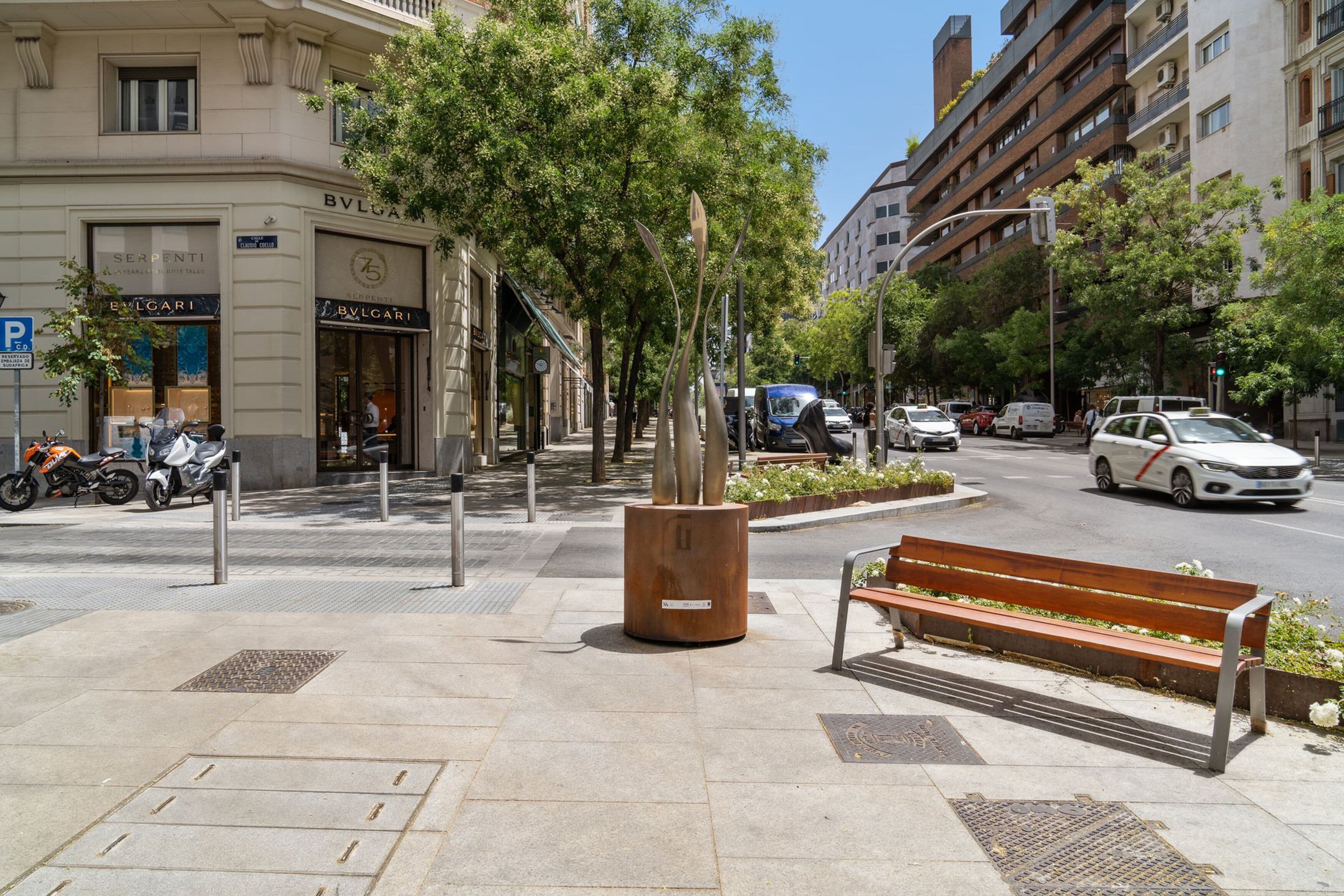 Una calle urbana con aceras, bancos y vehículos estacionados.